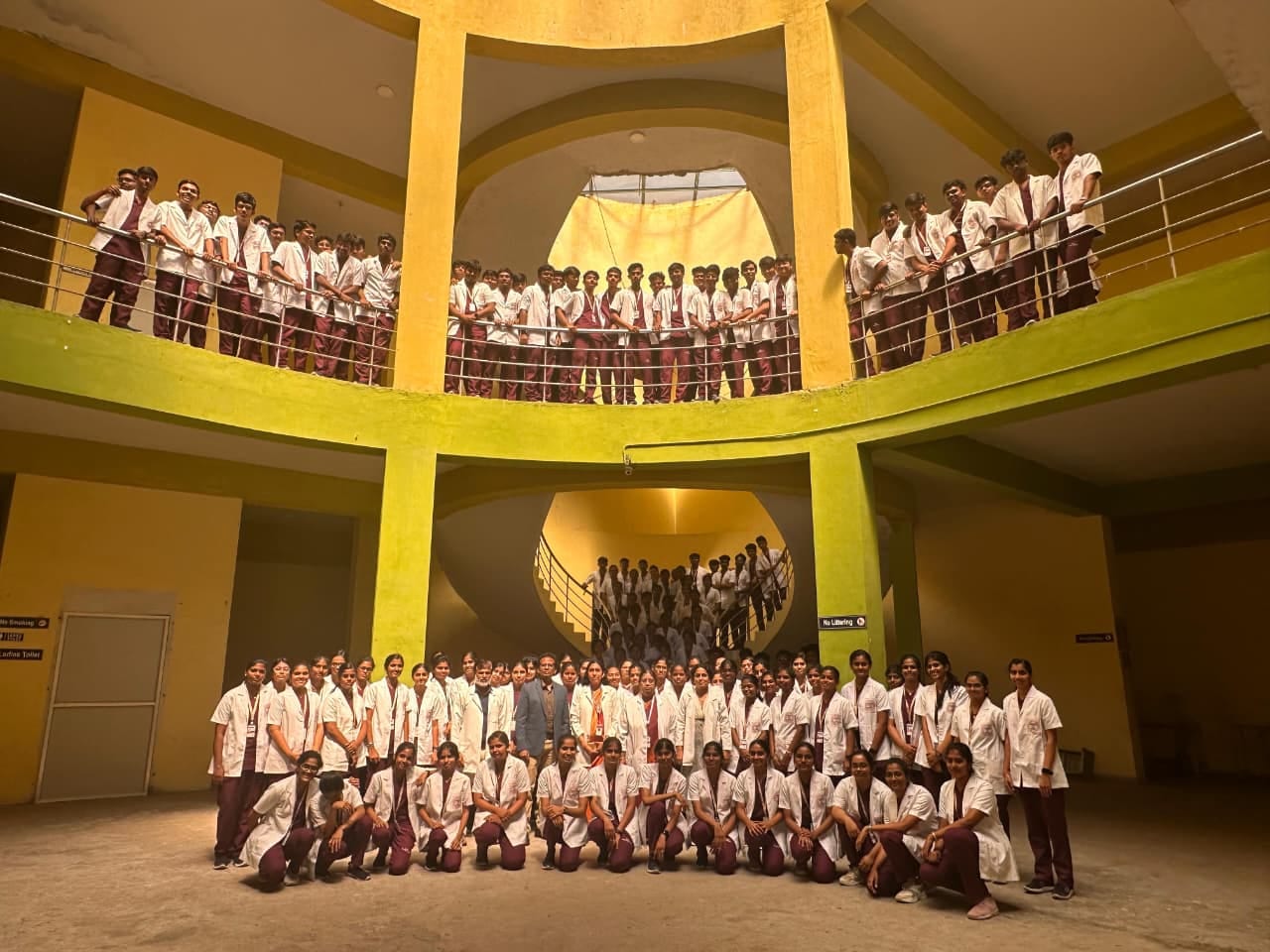 A large group of students in white coats and maroon scrubs gathered across a multi-level staircase and balcony for a group photograph.